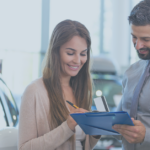 Man holding car contract on clipboard in front of vehicle with woman signing it | Increase Car Sales Despite Rising Costs With These 5 Tips | Safeguard